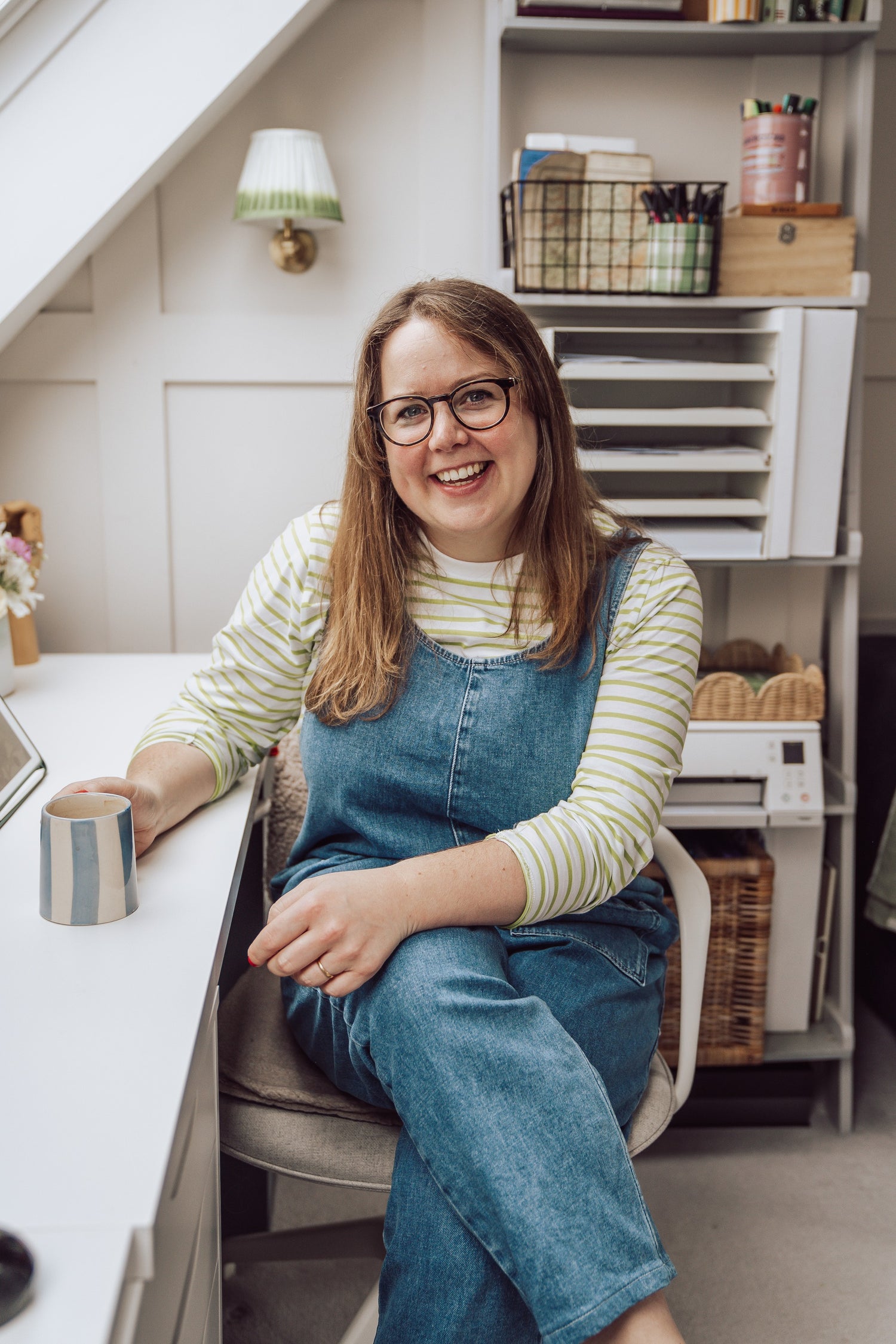 Portrait of Debbie Poulton, Surrey artist and illustrator sitting in her Surrey studio holding a cup of coffee sat at a desk